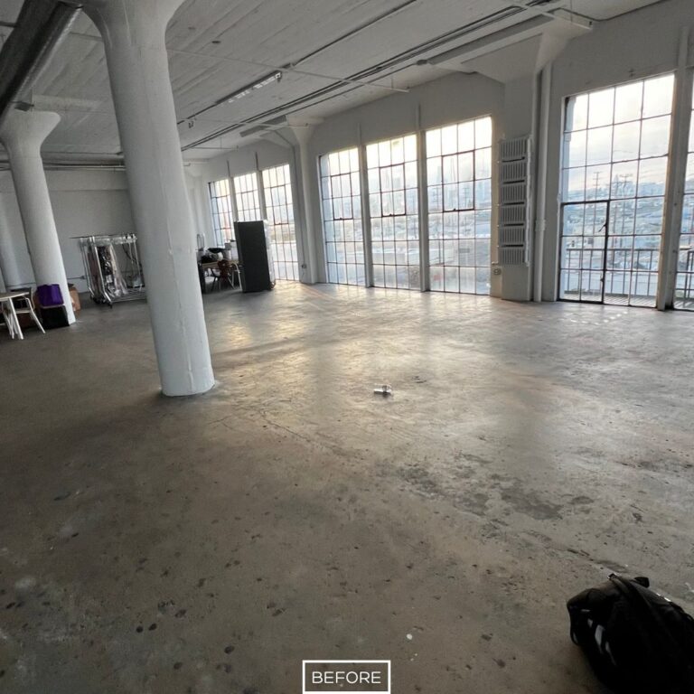 Unfinished concrete floor in Los Angeles loft before grinding, showing dull surface with uneven texture and stains near large windows.